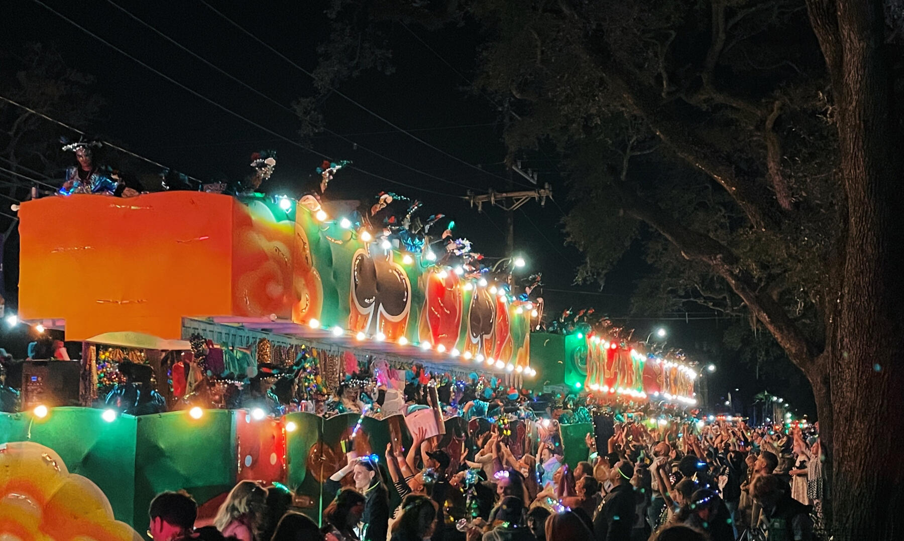 Looking down along the side of a log line of multicolored mardi gras floats at night among the crowd below arching oak trees.
